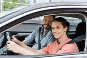 A father smiling in the passenger seat next to his teenage son behind the wheel of a car.